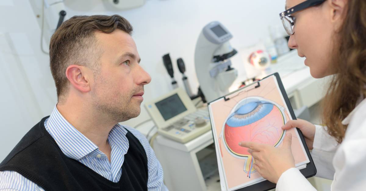 An ophthalmologist wearing a white lab coat uses an eye diagram to explain a health condition to her male patient.
