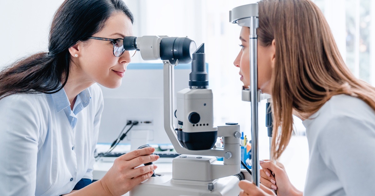 An eye care professional is examining a woman's eyes through a device, with her face resting on a headrest.