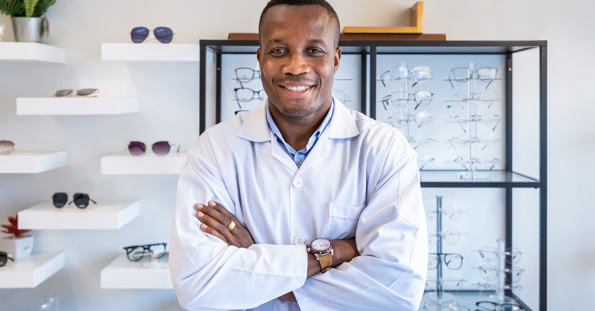 An optician in white garb is standing in front of a glass case of eye glasses, along with display shelves of sunglasses.