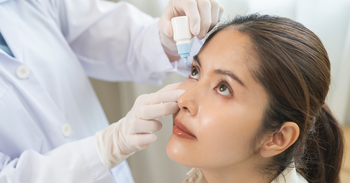 A woman is looking up while a medical professional in uniform with gloves puts eye drops in her eye.