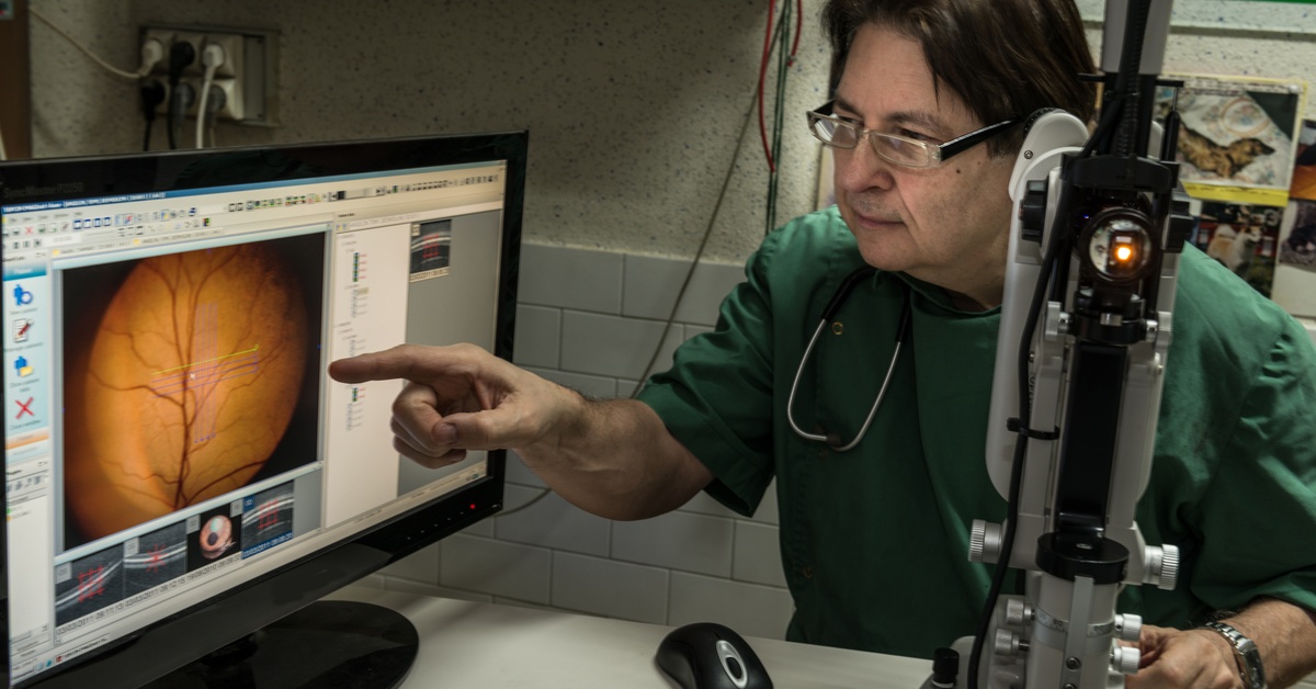 A medical professional is sitting in an office next to a tonometer. He is pointing at a screen displaying an eyeball.