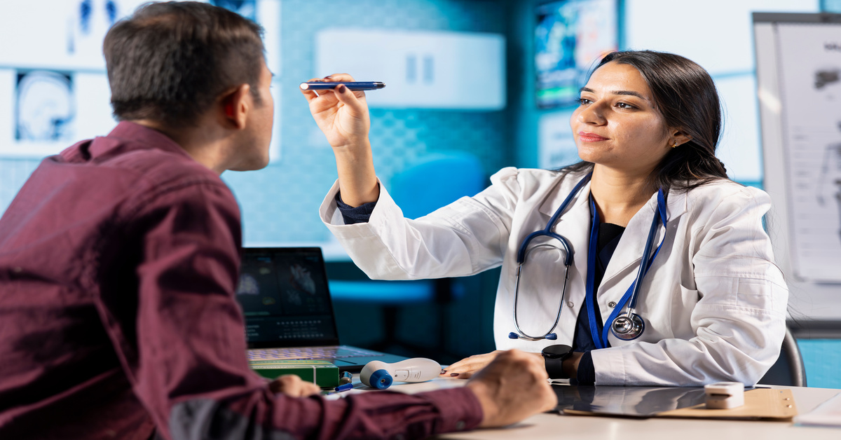 An ophthalmologist shines a penlight in a patient's eye in front of a desk containing a laptop and several medical supplies.