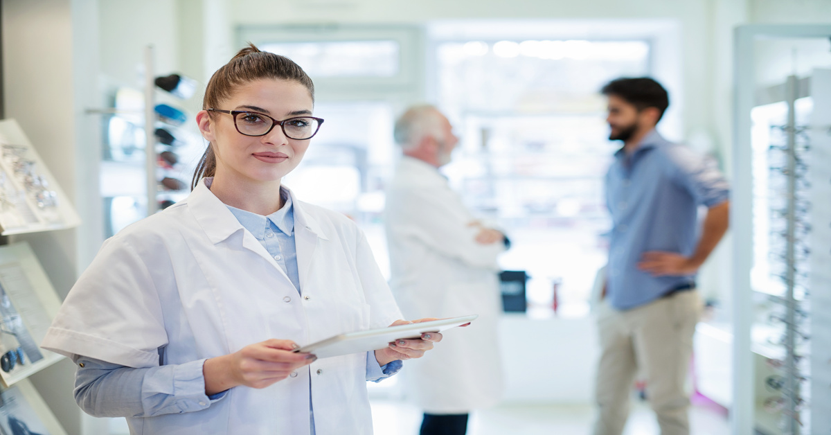 A woman in a white coat holds a tablet while standing in an optical shop with colleagues and glasses frames in the background.