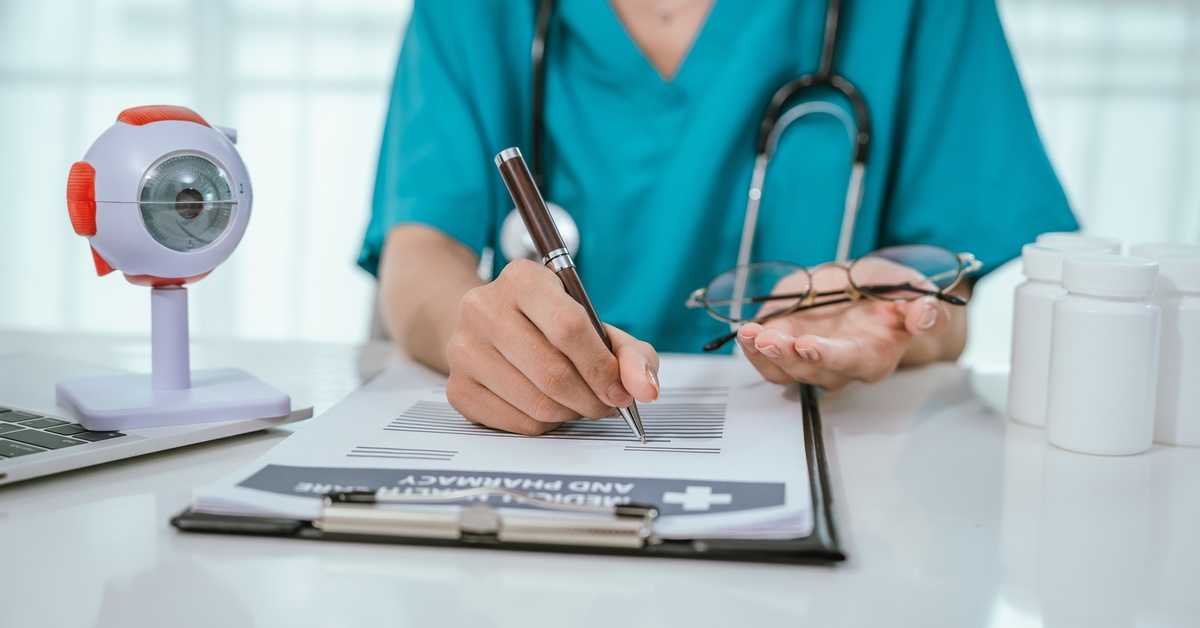 A person wearing scrubs sits at a desk. They're holding a pair of glasses in one hand and writing on a clipboard with the other.