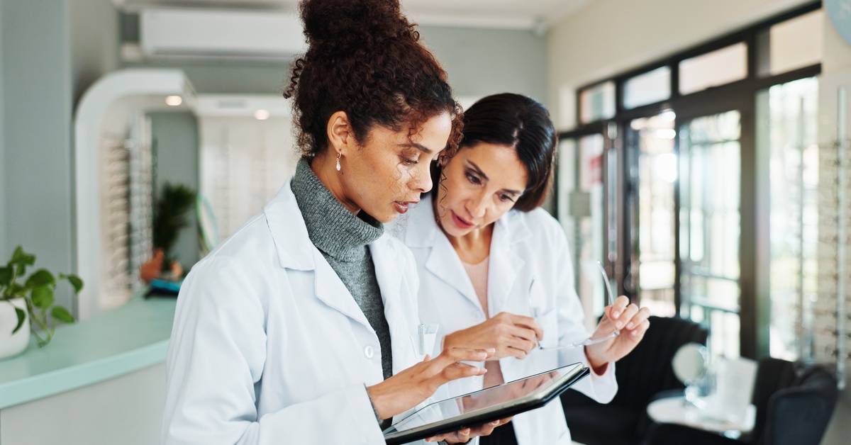 Two women wearing white lab coats look at a digital tablet that one of them is holding in a health clinic.