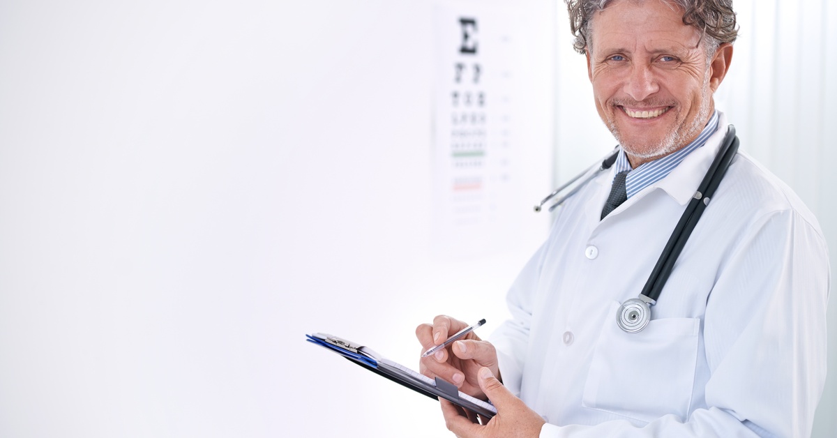A man in a white lab coat stands in front of a Snellen chart mounted on the wall, holding a clipboard and pen.