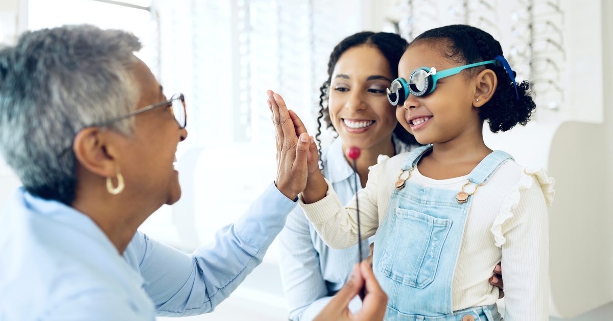 A woman holds a young girl wearing prescription eyeglasses as the child gives a high-five to a medical professional.