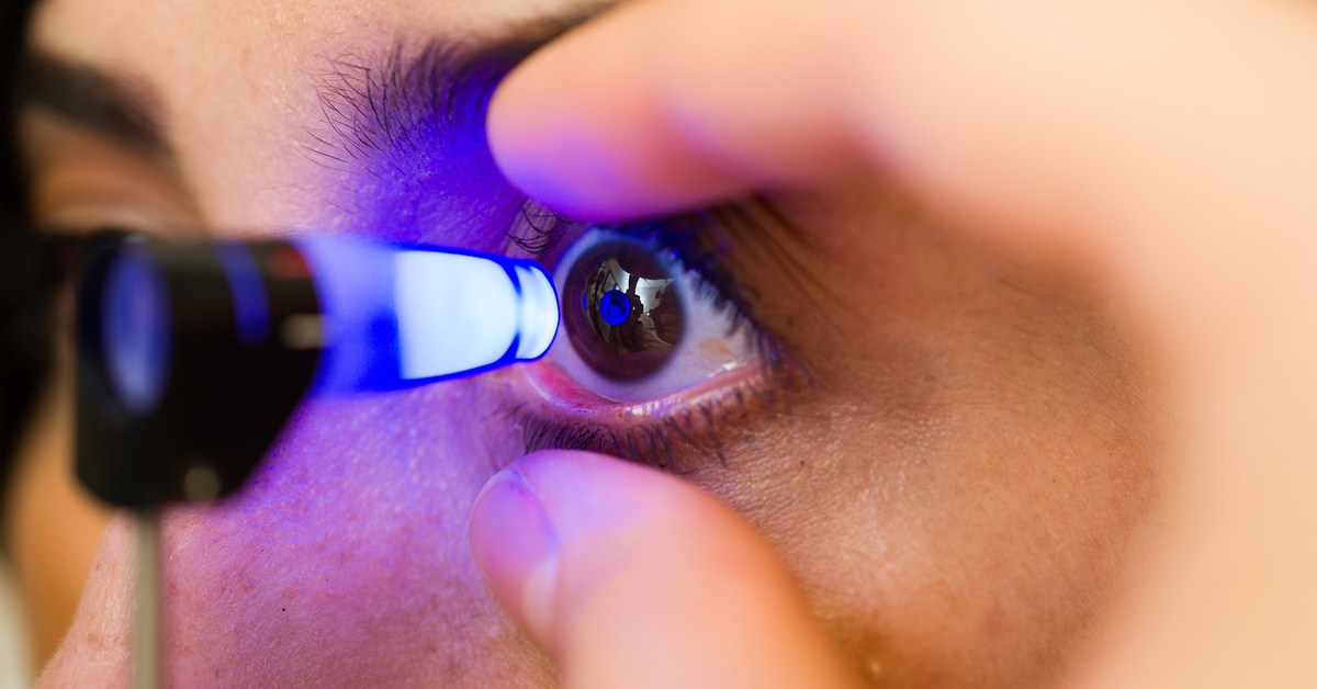 A person is looking into a small cone-like device that lights up blue, while a person holds their eyelids open.