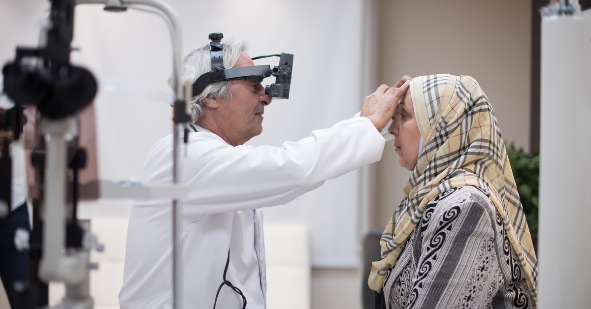 A woman wearing a hijab sits in front of a medical professional who performs an eye exam using professional tools.
