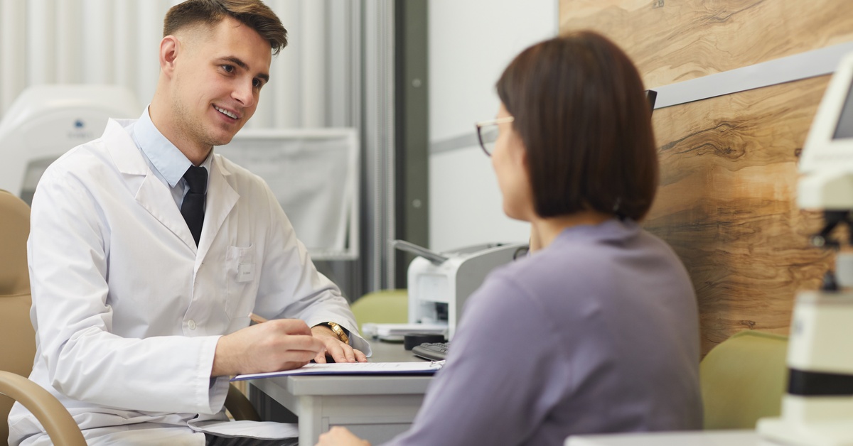 A woman in a white coat is talking with a seated woman near eye exam equipment in a bright clinic office.