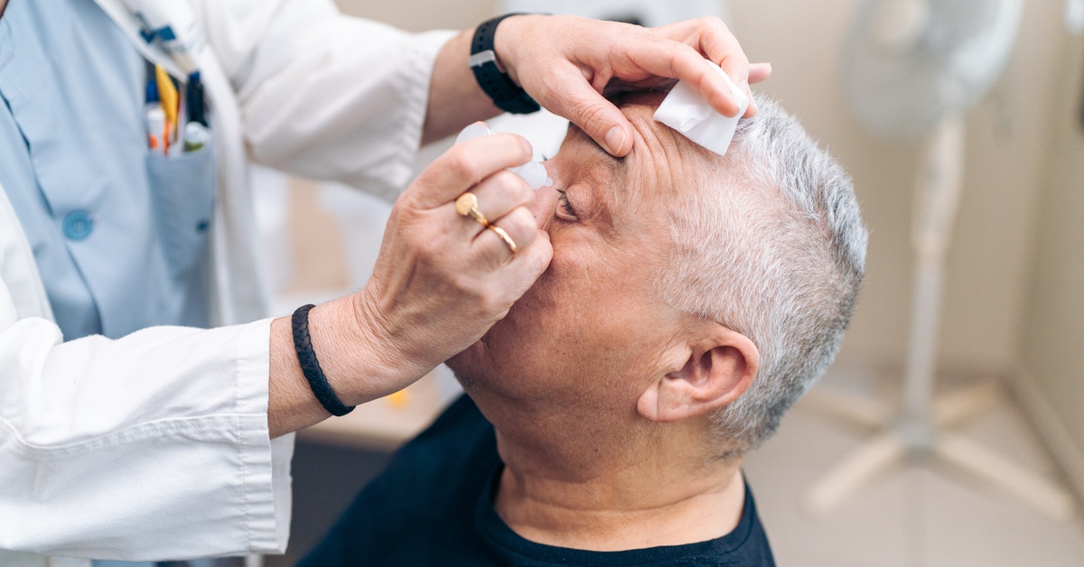 A senior man sits while a person in a lab coat uses alcohol wipes to clean his eyelids. A fan is visible in the background.