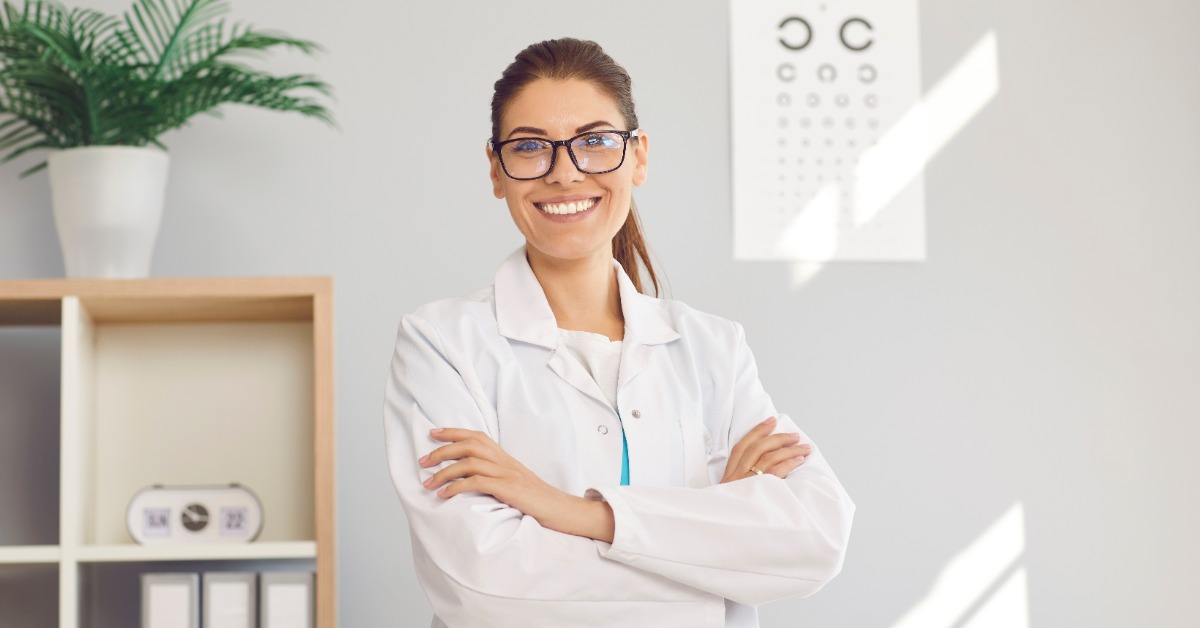 A woman wearing glasses and a white coat smiles with arms crossed, standing near shelves, a plant, and an eye chart.