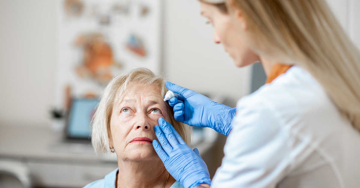 A woman wearing blue gloves applying eye drops to an older woman looking upward in a bright medical office.