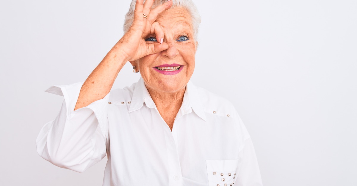 A gray-haired woman smiles and makes a hand gesture over one eye while standing next to a white background.