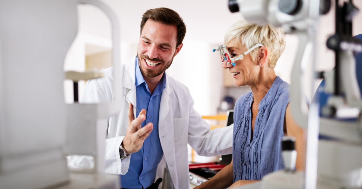 A man in a white coat speaks with a woman wearing trial frames, who sits between ophthalmic machines in a bright exam room.